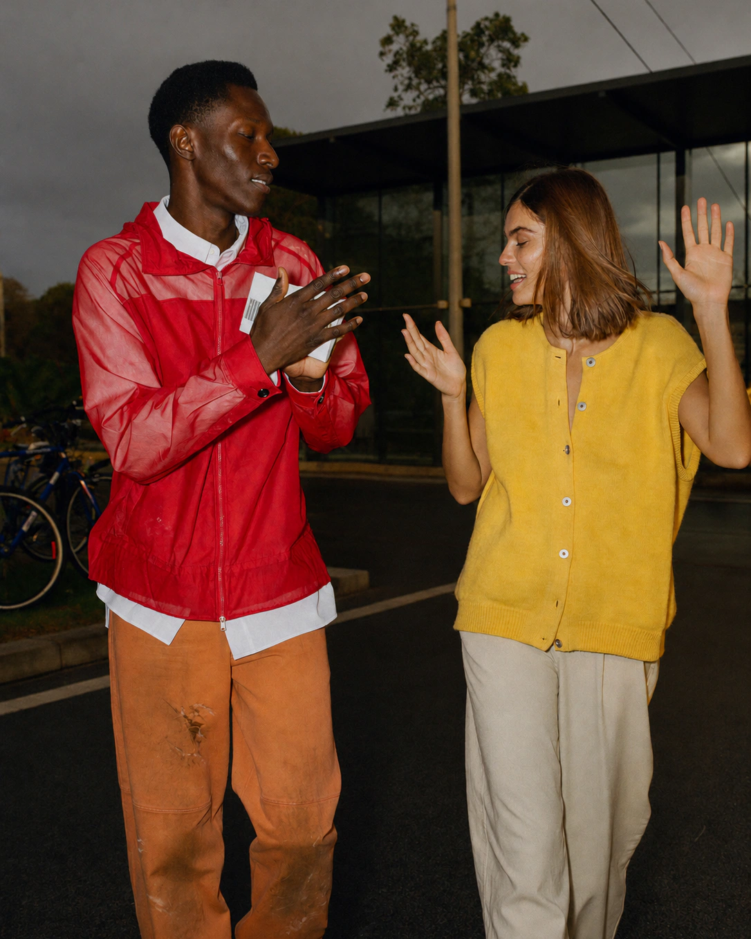 A man in a red zip jacket and a woman in a yellow sleeveless knit gesture to each other outside at dusk.