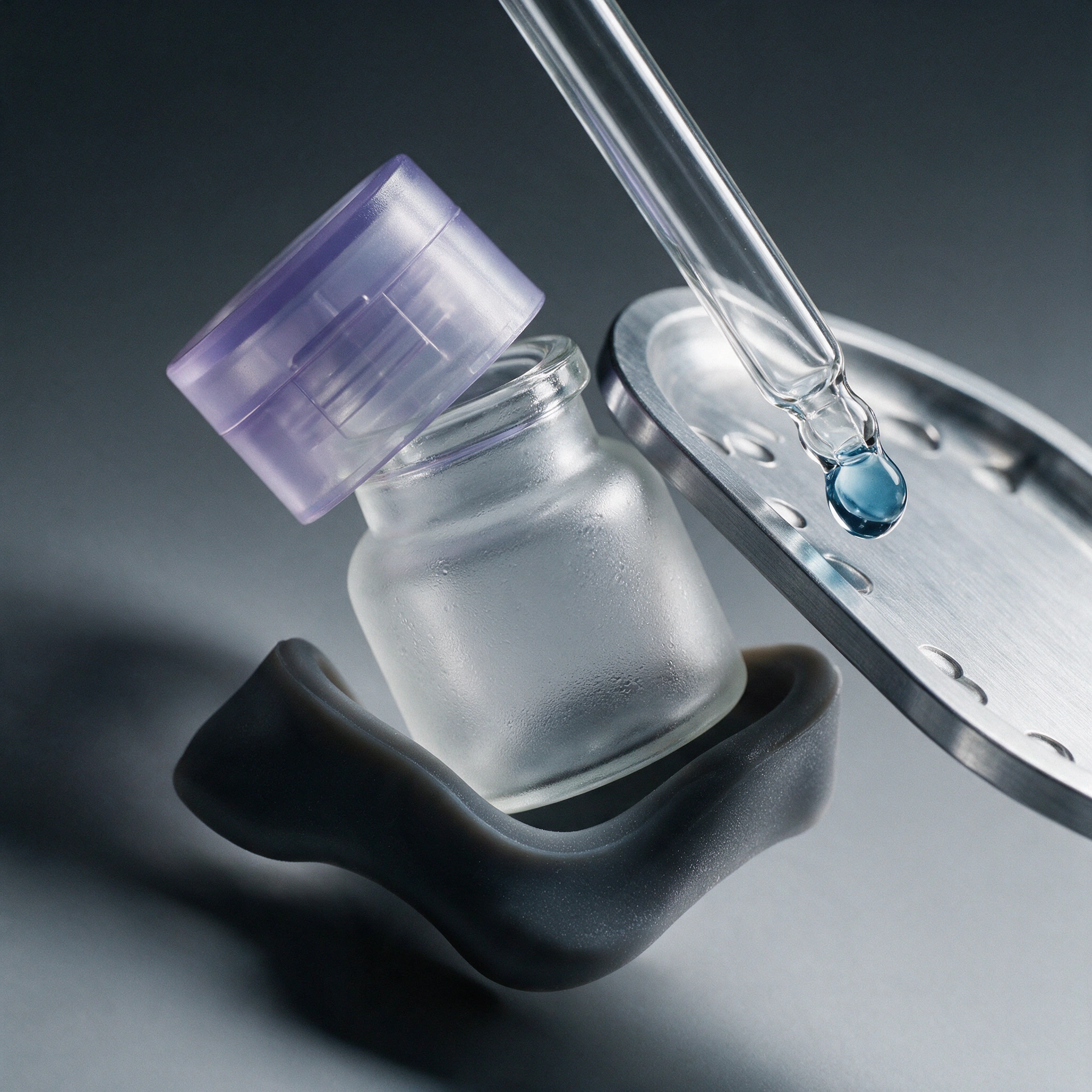 A frosted jar with a lilac cap beside a glass dropper holding a blue droplet, set in a dark holder beside a silver tray.