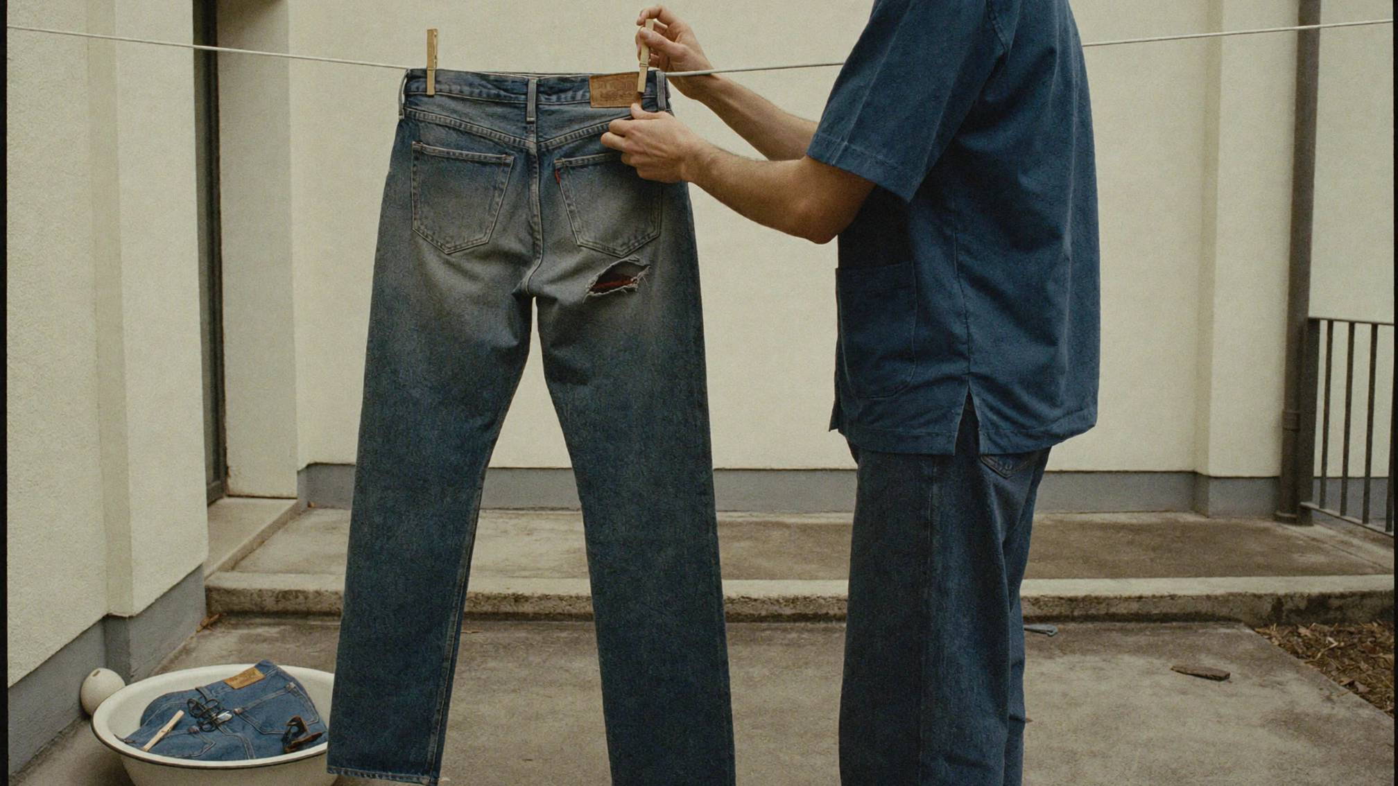 A person in a blue shirt clips a pair of faded jeans to a line in a courtyard, with a basin below.