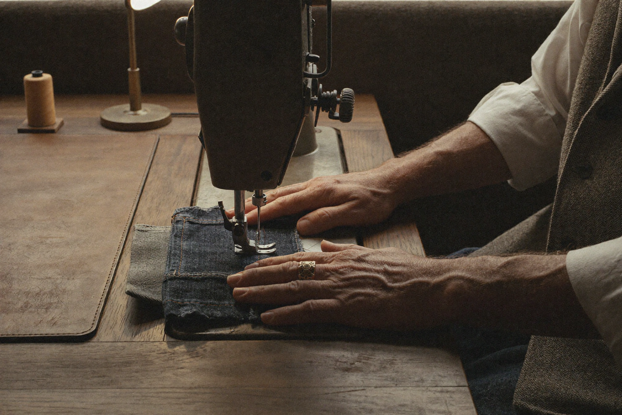 Hands guide a folded section of denim beneath a black sewing machine on a wooden table.