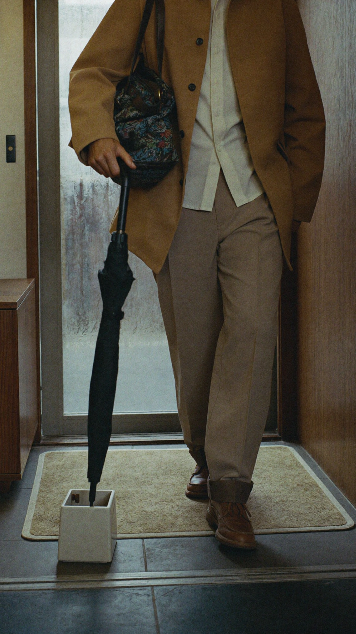A person in a tan suit holds a black umbrella beside a small pale stand at an apartment entrance.