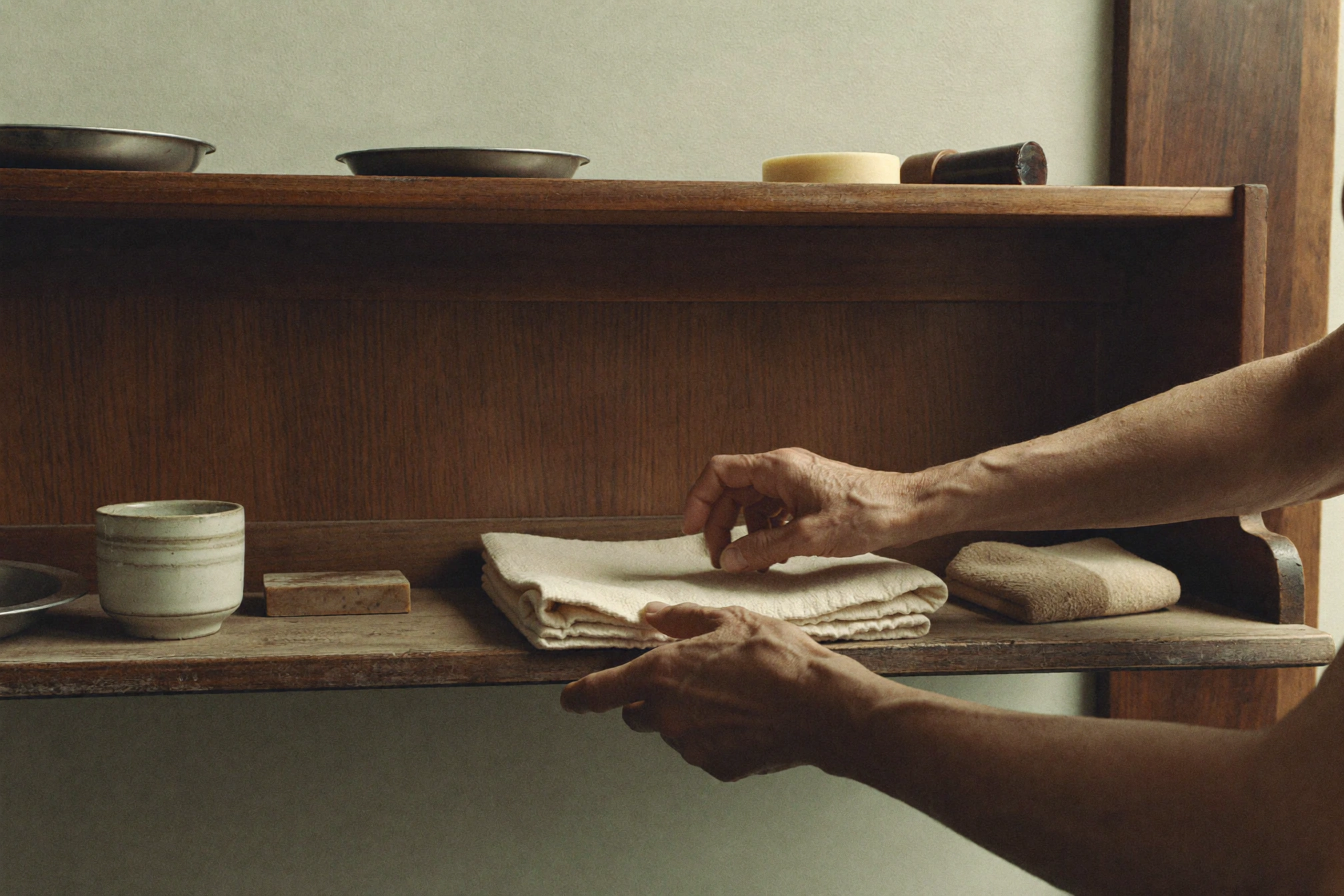 Two pairs of hands handle folded off-white cloths on a wooden shelf beneath cups, trays, and a small brush.