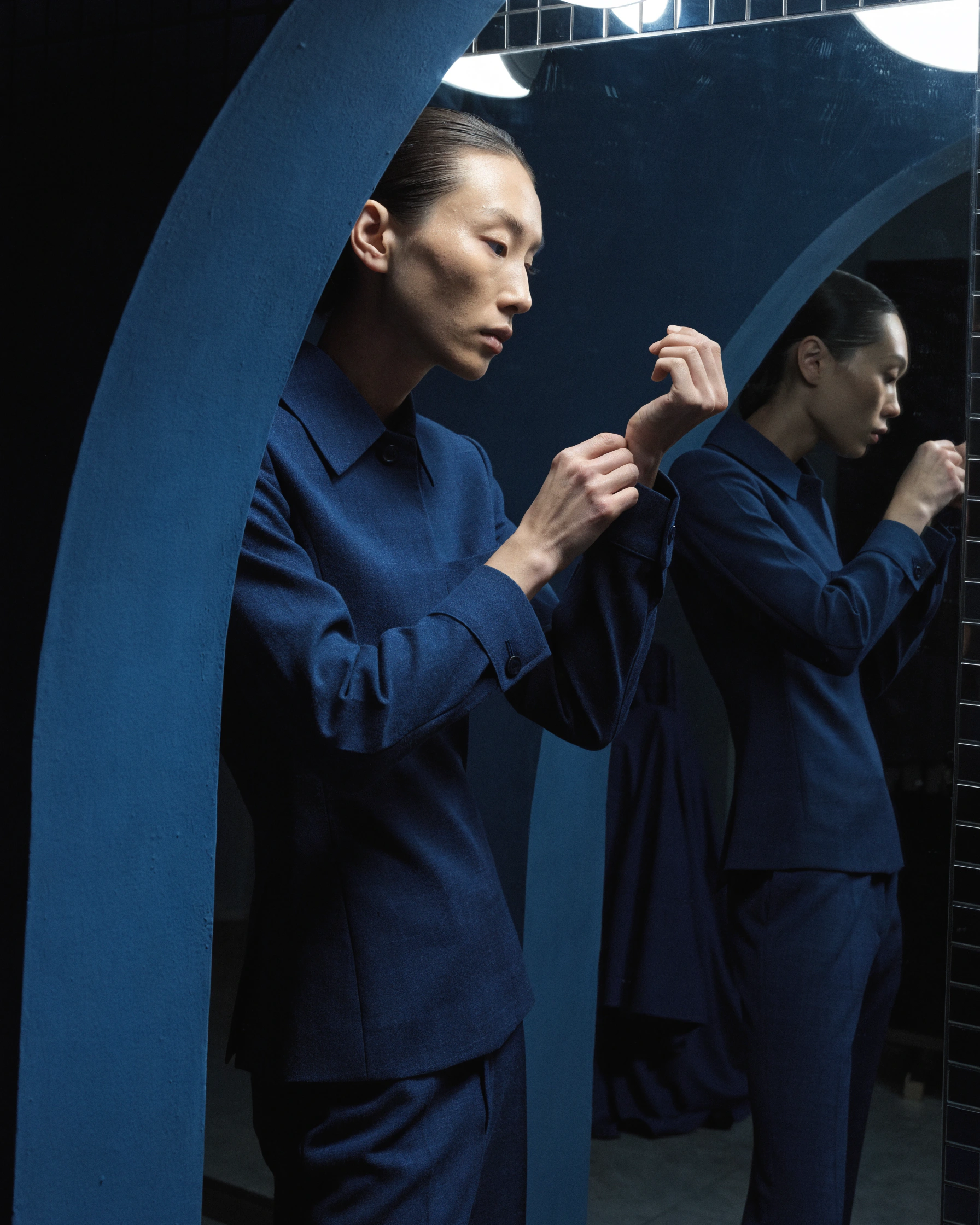 A model adjusts one cuff before a mirror lit only by cool work lights.