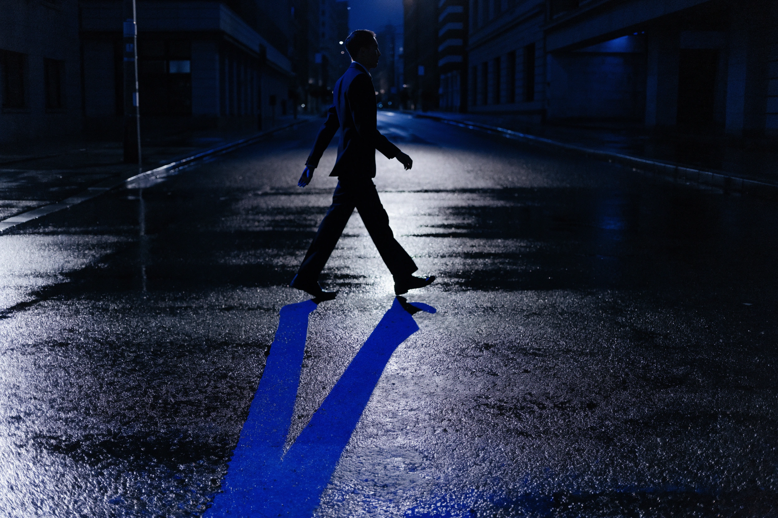 A single silhouette crosses an empty street between hard pools of blue light.