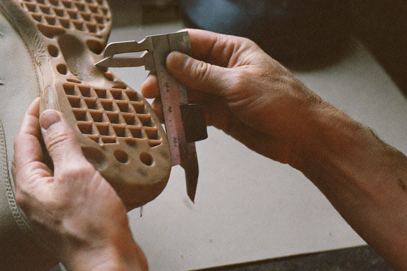 Hands hold a tan shoe while a caliper is placed against its raised outsole tread.
