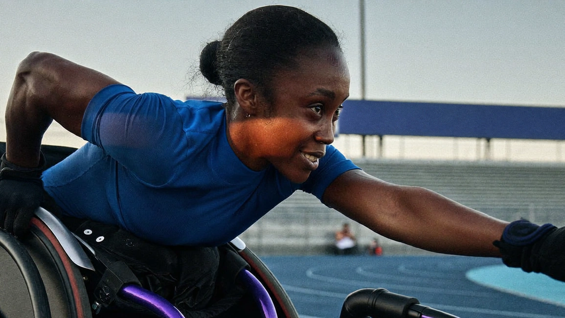 A wheelchair athlete in a blue shirt leans forward and reaches across an indoor blue sports surface.