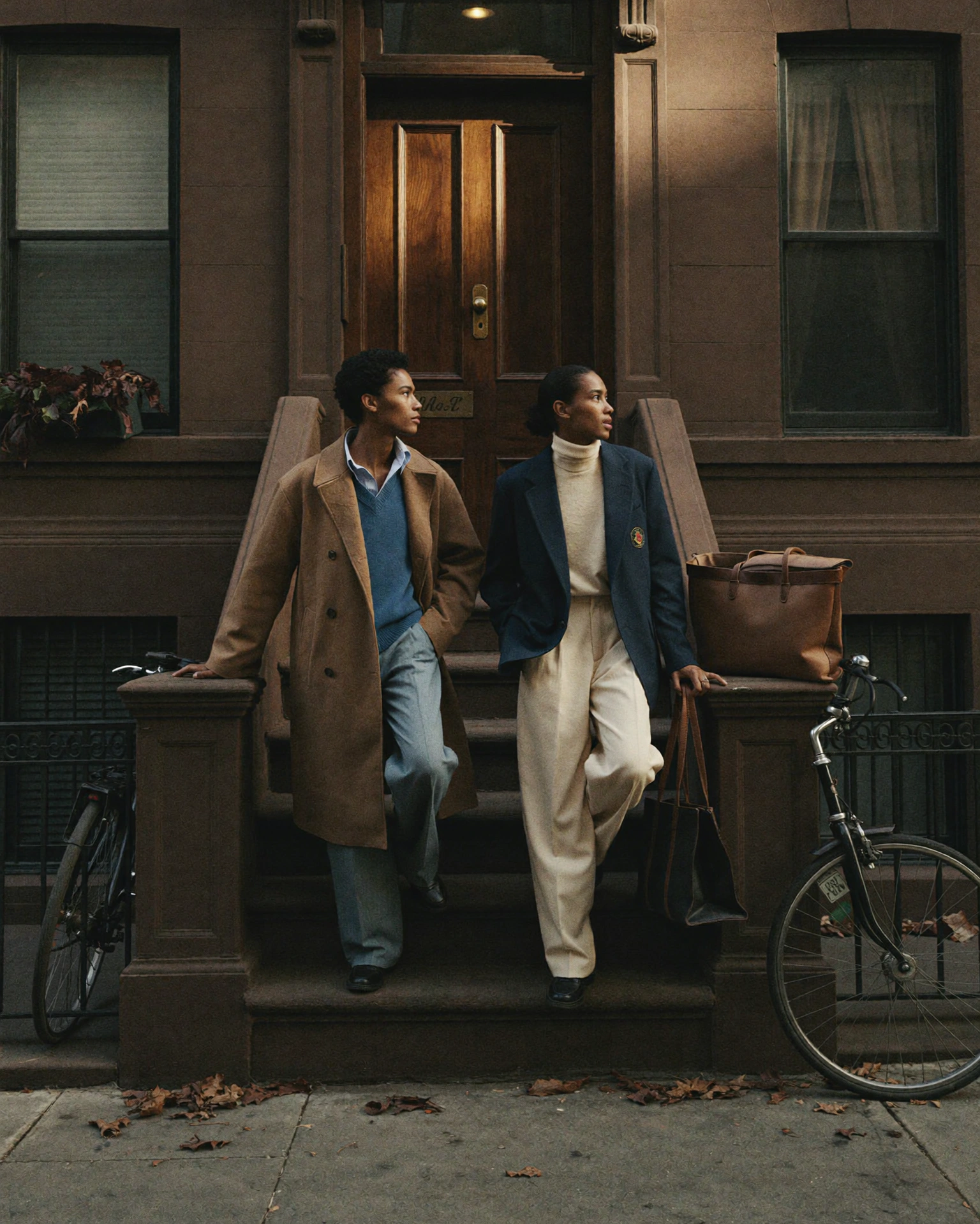 Two people stand on the steps of a brownstone entrance, framed by bicycles and a large brown tote.
