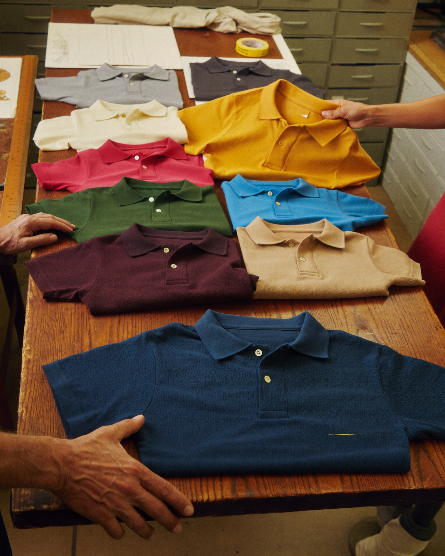 Hands arrange folded polo shirts in cream, navy, green, yellow, red, and blue across a table.
