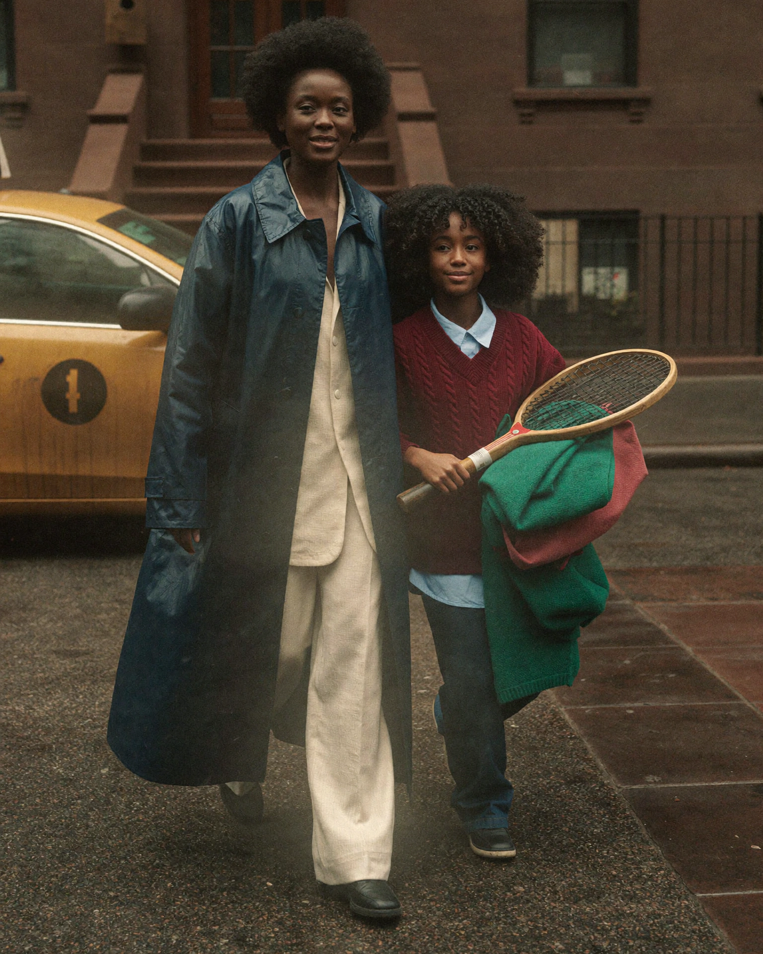 A woman in a navy raincoat and a child holding a wooden racquet walk on a wet city street beside a yellow car.