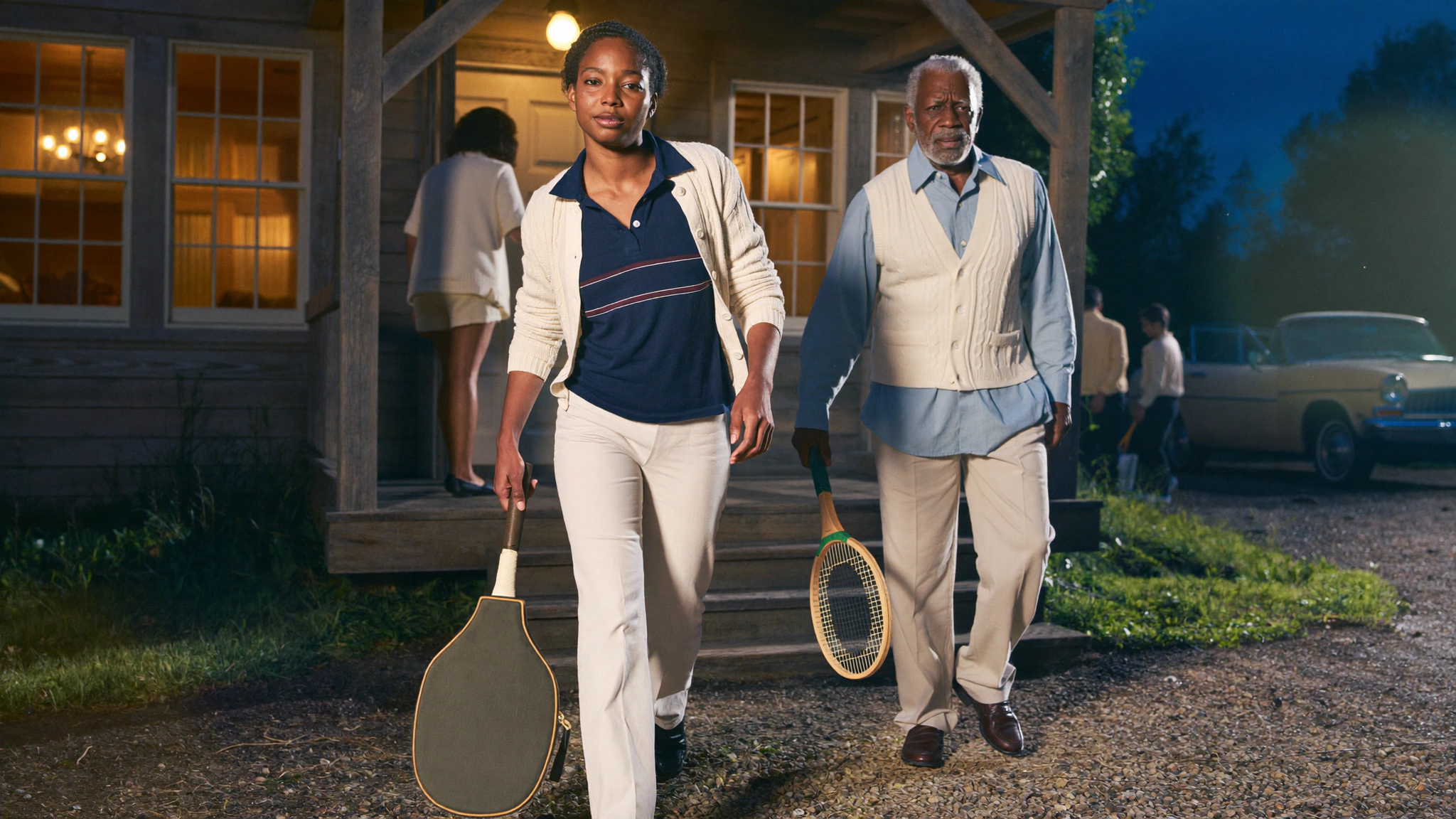 A woman and an older man walk toward the camera from a lit clubhouse at night, carrying a racquet case and an uncovered racquet.