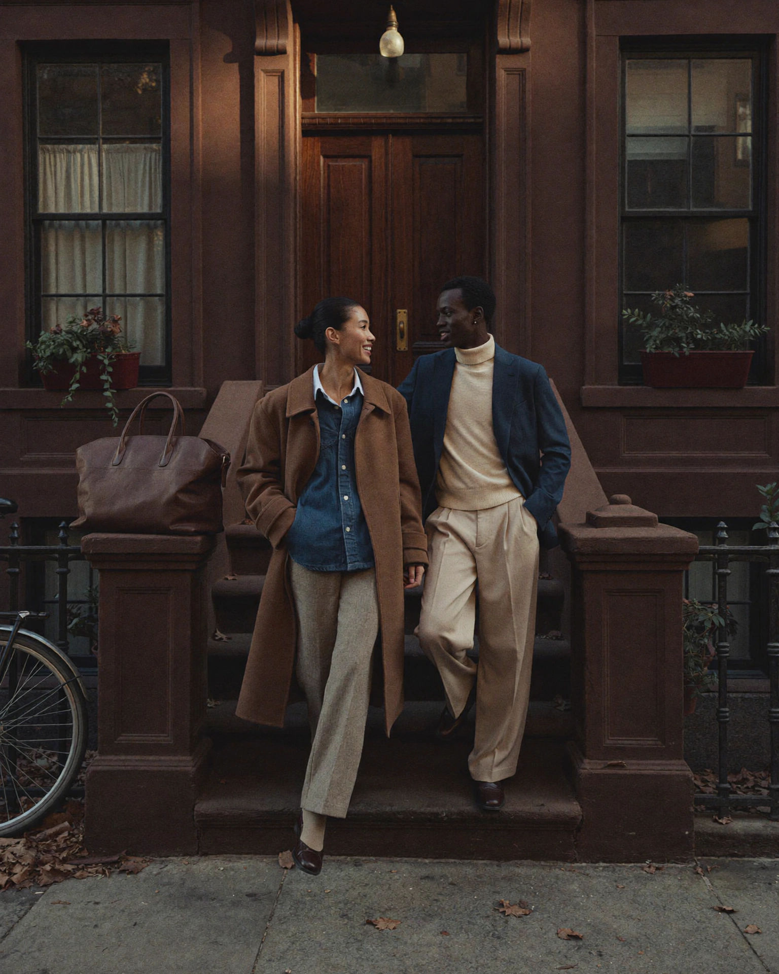 Two people in brown, cream and navy layers stand close together on a brownstone stoop, with a bicycle at the edge.