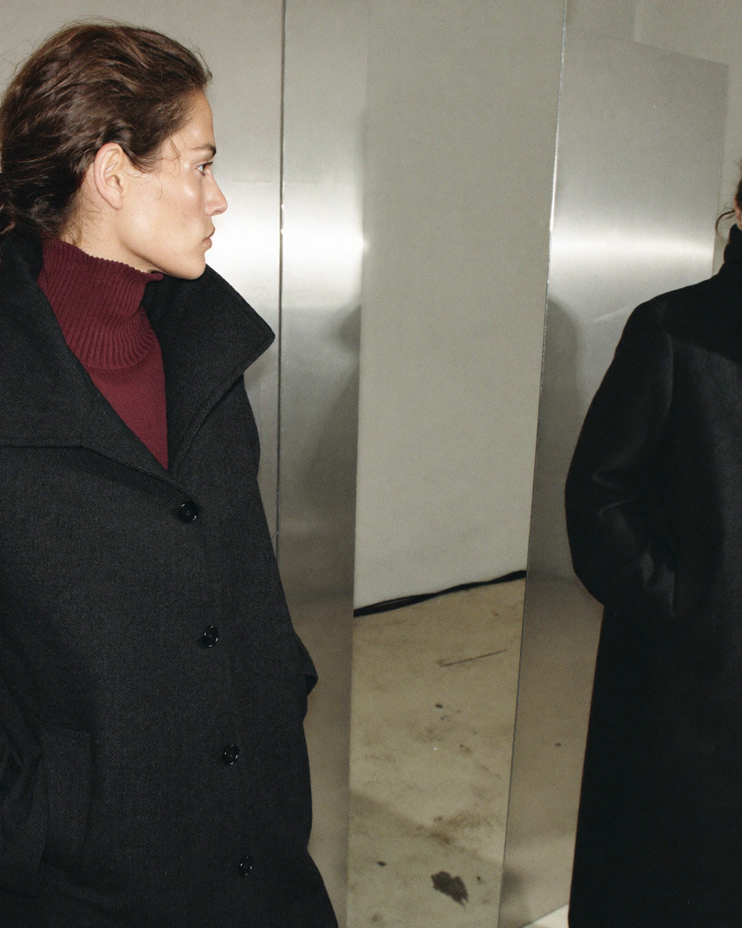 A woman in a black coat and burgundy knit stands beside her reflection in a mirror.