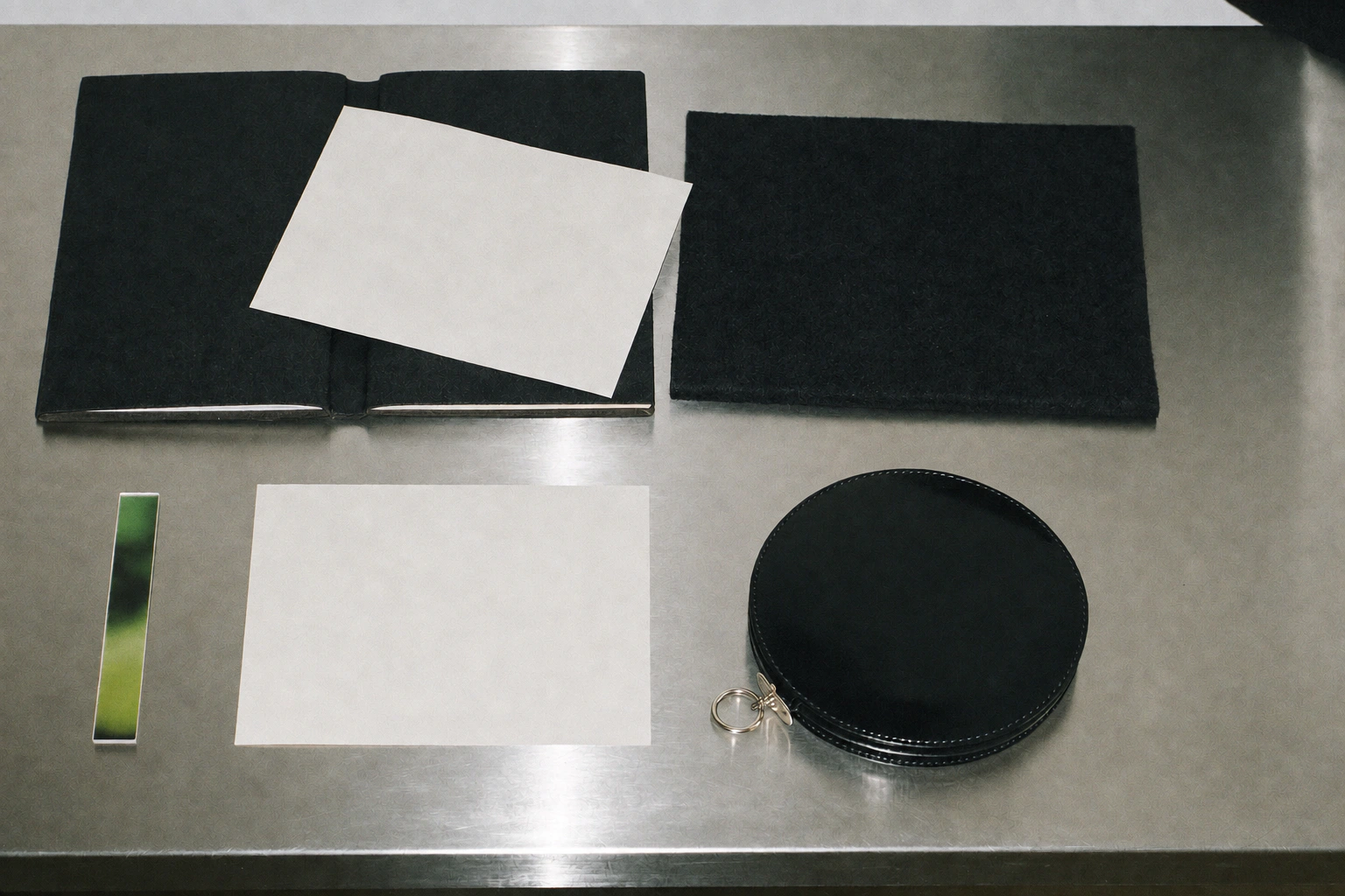 Black leather folios, white paper, a rounded leather object and a green metal strip lie on a reflective table.