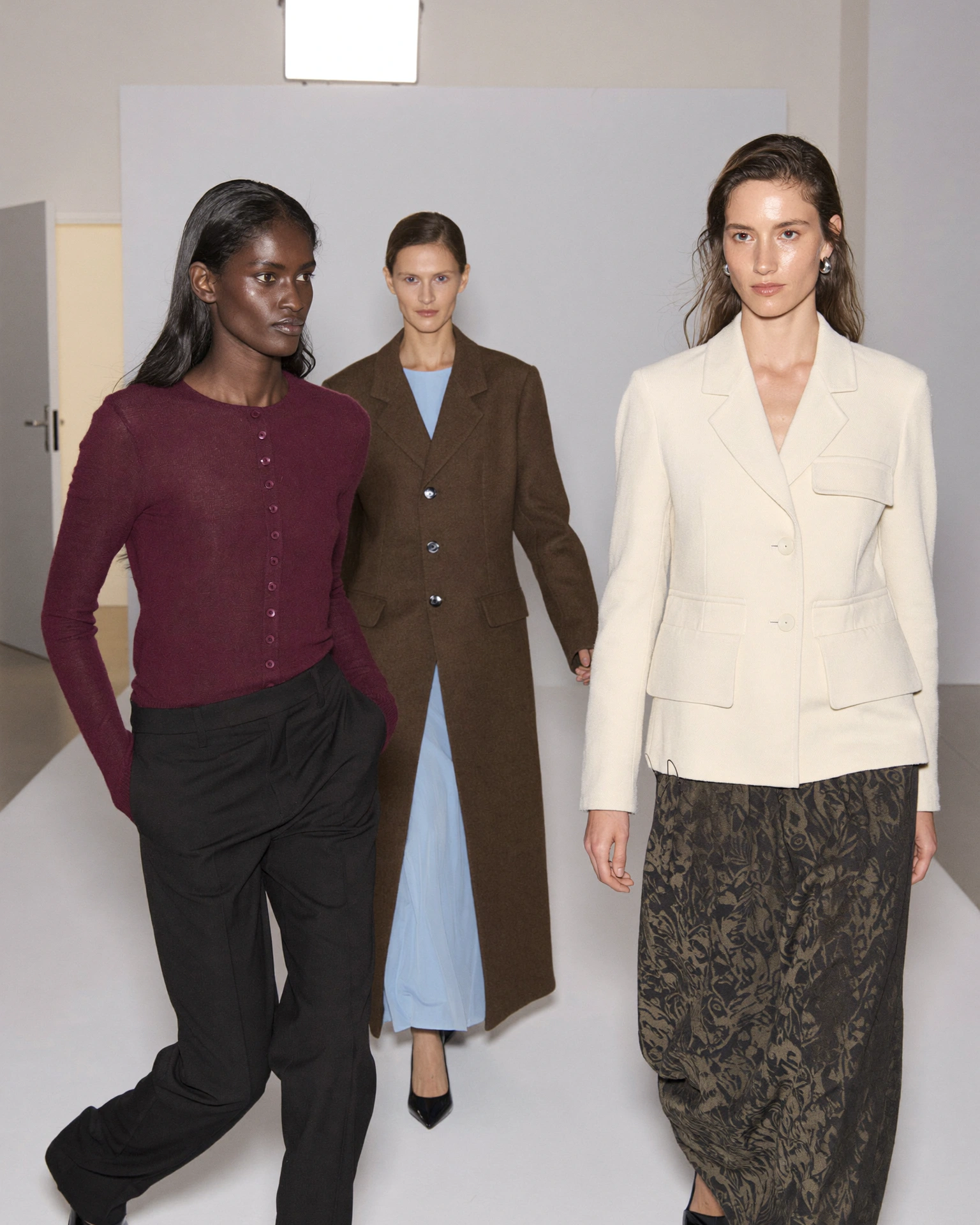 Three models in burgundy, brown, pale blue and ivory clothes stand together in a white casting room.