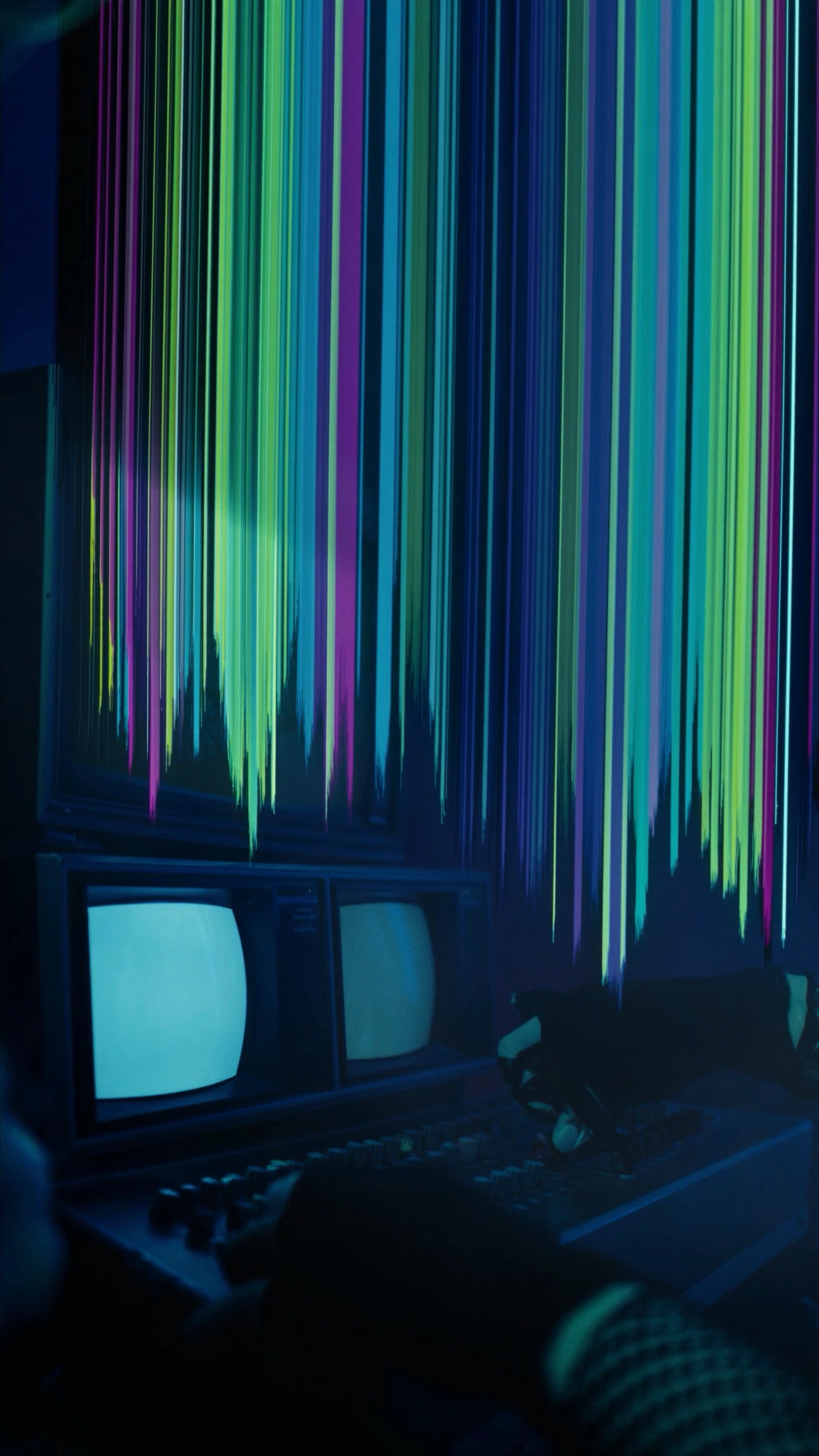 Hands operating an analog video mixer before a wall of colour-shifted CRT monitors.
