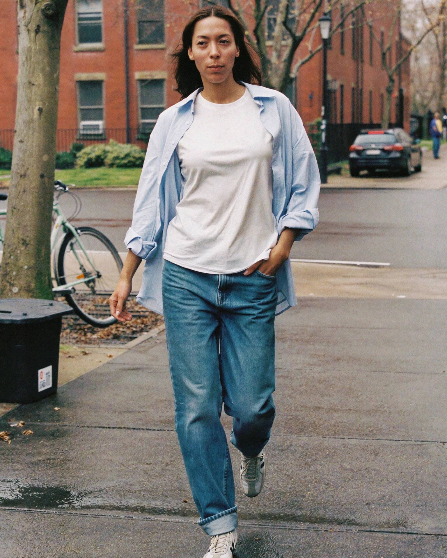 A woman wearing a white T-shirt, open light blue shirt and jeans walks through a brick-lined neighbourhood.