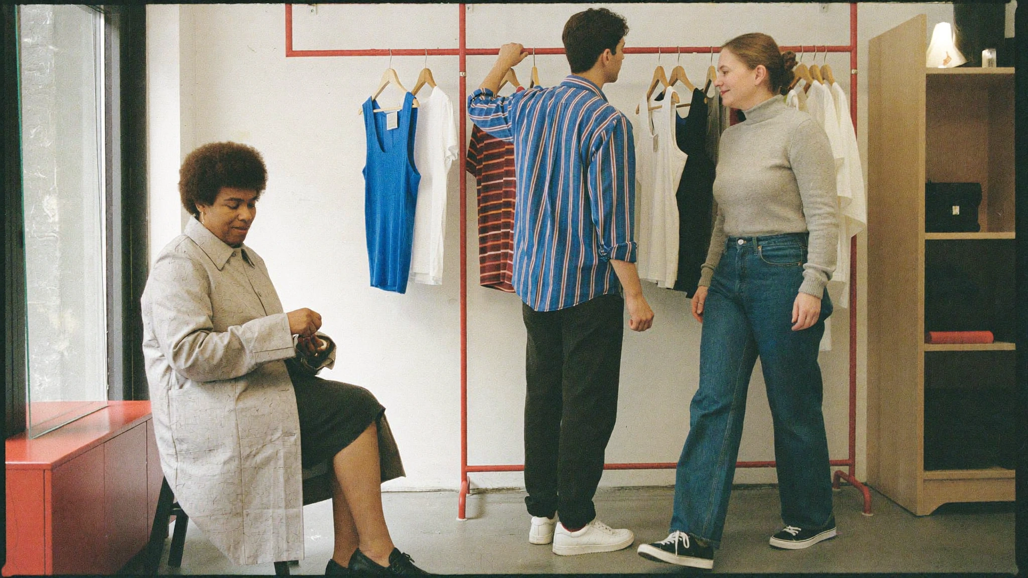 Three people choose and wear simple layered clothes in a bright room with a red clothing rail and wooden shelving.