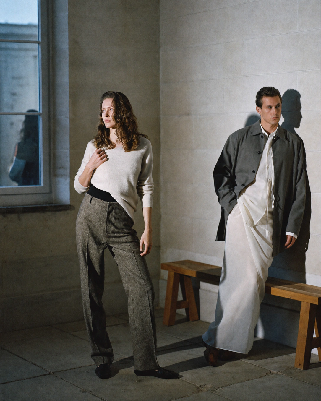 Two people in grey tailoring and cream knitwear stand apart in a sunlit stone room.
