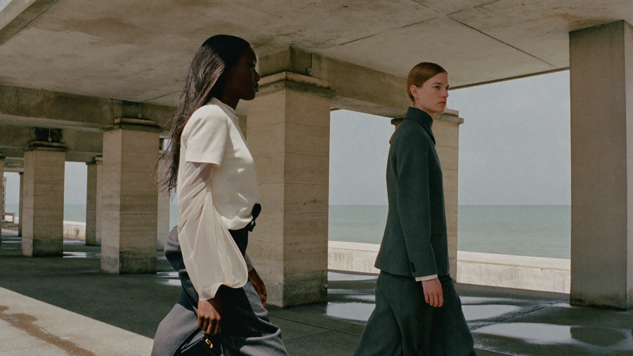 Two models in white and charcoal walk past concrete columns beside the sea in directional afternoon light.