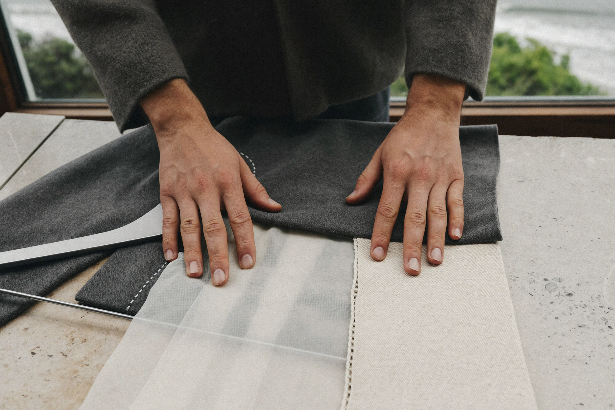 Hands arrange charcoal wool, translucent white cloth and cream fabric on a stone table beside a window.